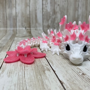 May include: A white and pink 3D printed dragon figurine with a flower-shaped tail. The dragon has pink flowers on its head and back.