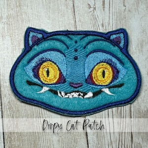 May include: A teal embroidered patch featuring a cartoon cat face with large yellow eyes, purple accents, and a toothy grin. The patch has a dark blue border and the text "Dirpy Cat Patch" is below the cat's face.