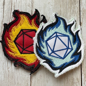 May include: Two embroidered patches featuring a polyhedral die design. One patch has a red die surrounded by yellow, orange, and black flames. The other has a white die with blue flames and a light green background. Both patches have a black border.