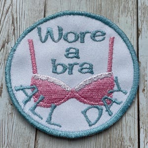 May include: Embroidered patch with a pink bra design and the words "Wore a bra ALL DAY" in light blue. The patch is circular, with a white background and a light blue border.