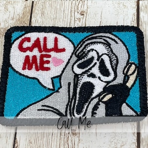May include: Embroidered patch featuring a gray figure with a white mask holding a phone. The speech bubble says "CALL ME" in red letters with a pink heart. The patch has a black border and a teal background.