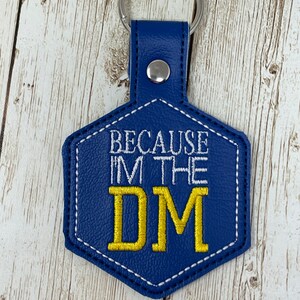 May include: A blue keychain with the text "BECAUSE I'M THE DM" in white and gold lettering. The keychain has a silver ring and a blue leather-like tag in a hexagonal shape.