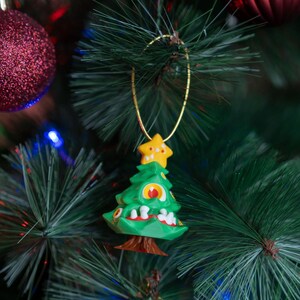 May include: A green Christmas tree ornament with a yellow star on top and a white, red, and blue face. The ornament is hanging on a green Christmas tree.