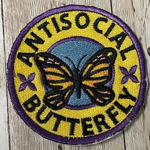 Puede incluir: Un parche redondo bordado de color amarillo con un borde morado. El parche presenta una mariposa negra con detalles amarillos y rosados, y el texto "Antisocial Butterfly" en negro.