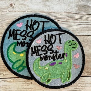 May include: Two embroidered iron-on patches with a green dinosaur and the text "Not Mess Monster" on a light blue background.