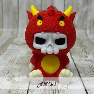May include: A red devil-like figurine with yellow horns and eyes, and a white skull-shaped face. The figurine has a yellow circle on its chest and the word "Scorchi" is written at the bottom.