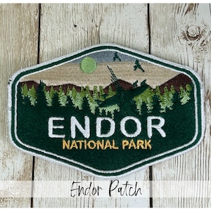 May include: An embroidered patch featuring a landscape scene with a green sun, dark green forest, and brown mountains. The text ENDOR NATIONAL PARK is stitched in white and yellow thread. The patch has a white border and a hexagonal shape.