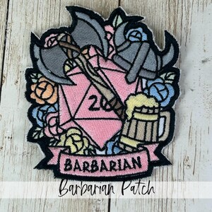 May include: Embroidered patch featuring a pink D20 die with the number 20, crossed axes, and a mug of beer. The patch is surrounded by roses in shades of pink, blue, and yellow, with the word "BARBARIAN" on a banner.