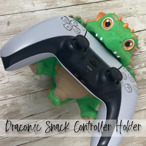 May include: A green dragon-shaped controller holder with orange accents and large eyes, holding a white and black wireless game controller. The base of the holder features the text Draconic Snack Controller Holder in black script lettering.