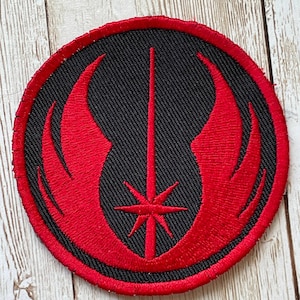 May include: Round black patch with a red emblem featuring a central starburst and stylized wings. The patch has a red border and is likely an embroidered design, suitable for application on clothing or accessories.
