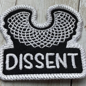 May include: Black and white embroidered patch with a white outline. The patch features a white stylized image of a judge's collar and the word "DISSENT" in white lettering.