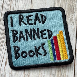 May include: A blue embroidered patch with black text that reads "I read banned books" and a stack of colorful books on the right side.