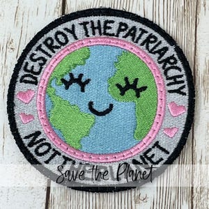 May include: Embroidered patch with a circular design. The patch features a smiling Earth with closed eyes, surrounded by the text "DESTROY THE PATRIARCHY NOT THE PLANET." Pink hearts and a pink border encircle the Earth. The words "Save the Planet" are written below.