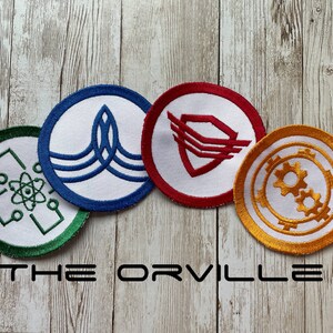 May include: Four embroidered patches with circular designs in green, blue, red, and gold. Each patch features a unique emblem, set against a white background. The text "THE ORVILLE" is displayed in black block letters below the patches.
