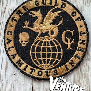 May include: Embroidered patch featuring a black background with gold text and a gold dragon design. The text reads "THE GUILD OF CALAMITOUS INTENT". A skull and globe are also featured in the design. The Venture Bros. logo is in the bottom right corner.
