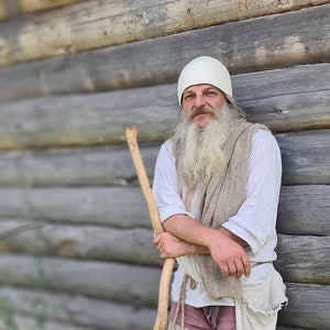 May include: A man with a long white beard and a white hat stands next to a wooden wall. He is wearing a white shirt, a beige shawl, and pink pants. He is holding a long, curved stick.