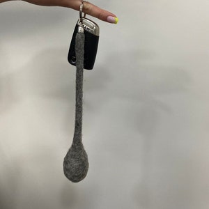 Könnte beinhalten: Ein grauer Filz-Schlüsselanhänger mit einem schwarzen Autoschlüssel. Der Schlüsselanhänger ist kugelförmig und hat eine lange, dünne Schnur.