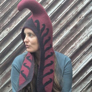 May include: A deep burgundy felted hood featuring a tall, curved top section and long, pointed side panels. The hood is decorated with a black, fern-like leaf pattern that runs along the edges and down the length of the side flaps.
