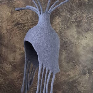 May include: A unique, light gray felt hat with a rounded crown and long, fringed edges. The hat has multiple, slender, tentacle-like protrusions extending from the top. The hat is suspended against a textured, brown and gray background.