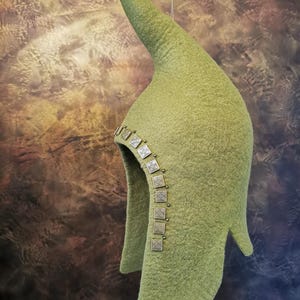 Può includere: Un cappello di feltro verde con una cima a punta e una fascia decorativa di quadrati di metallo argentato. Il cappello è appeso a una corda.