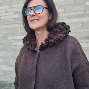 May include: A brown coat with a unique floral collar. The coat has a textured appearance and a button closure. The person in the image is wearing black-framed glasses.