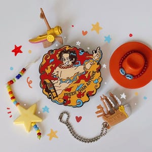 Peut inclure: Divers accessoires sur le thème des animés. L'image présente une épingle avec un personnage en flammes et le texte "Freer Than Anyone", une perle en forme d'étoile jaune, un petit navire et un chapeau orange avec des accents bleus.