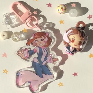 Peut inclure: Ensemble de porte-clés et de breloques sur le thème des animés. Les porte-clés comprennent un fermoir rose, des perles transparentes et un charm en acrylique transparent d'un personnage en tenue bleue et blanche. Une petite figurine et des perles décoratives sont également présentes, avec des confettis en forme d'étoiles.