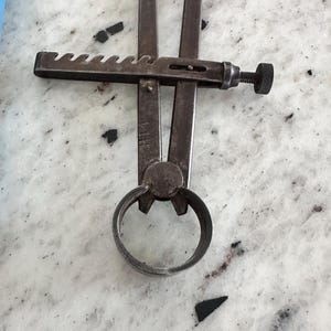 May include: A vintage metal caliper tool featuring two long, straight arms connected by a central pivot point. The tool includes a horizontal notched adjustment bar with a knurled screw mechanism for precise measurement settings and a circular ring base.