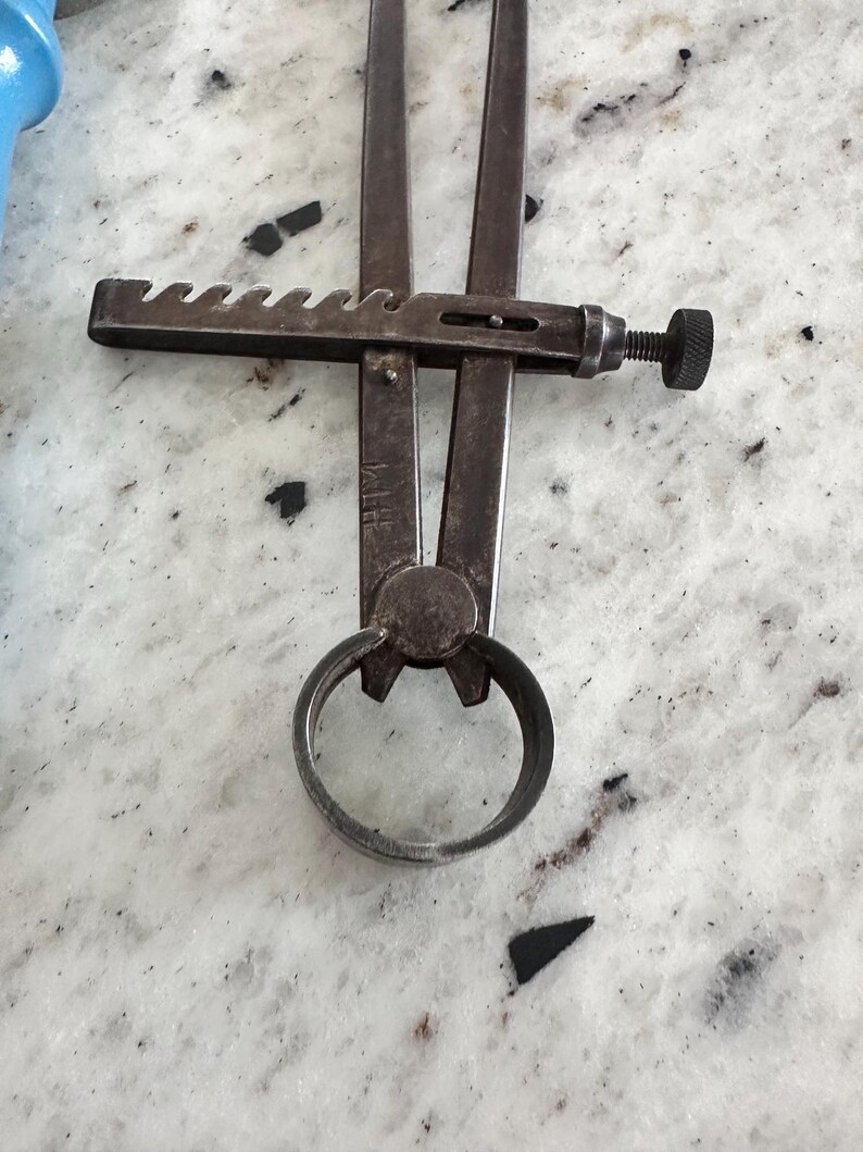 May include: A vintage metal caliper tool featuring two long, straight arms connected by a central pivot point. The tool includes a horizontal notched adjustment bar with a knurled screw mechanism for precise measurement settings and a circular ring base.