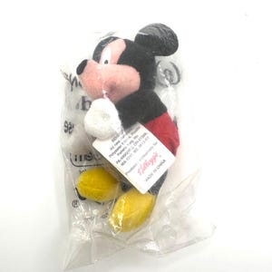 Pu&ograve; includere: Un piccolo peluche di Topolino in un sacchetto di plastica trasparente. Il giocattolo ha una testa e orecchie nere, una faccia rosa, pantaloncini rossi, scarpe gialle e guanti bianchi. Un'etichetta &egrave; attaccata al giocattolo e il testo "MADE IN CHINA" &egrave; visibile.