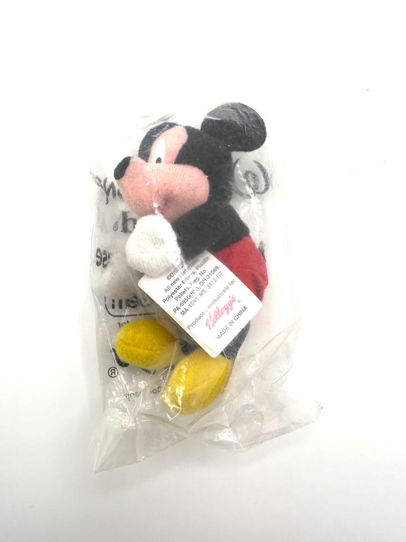 Pu&ograve; includere: Un piccolo peluche di Topolino in un sacchetto di plastica trasparente. Il giocattolo ha una testa e orecchie nere, una faccia rosa, pantaloncini rossi, scarpe gialle e guanti bianchi. Un'etichetta &egrave; attaccata al giocattolo e il testo "MADE IN CHINA" &egrave; visibile.