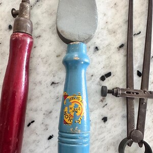 May include: A set of three tools including a red-handled awl, a blue-handled sharpening stone with a label reading Cyclone No. 272 A, and a metal caliper. The tools feature worn wooden handles and metallic components, showing signs of age and use.