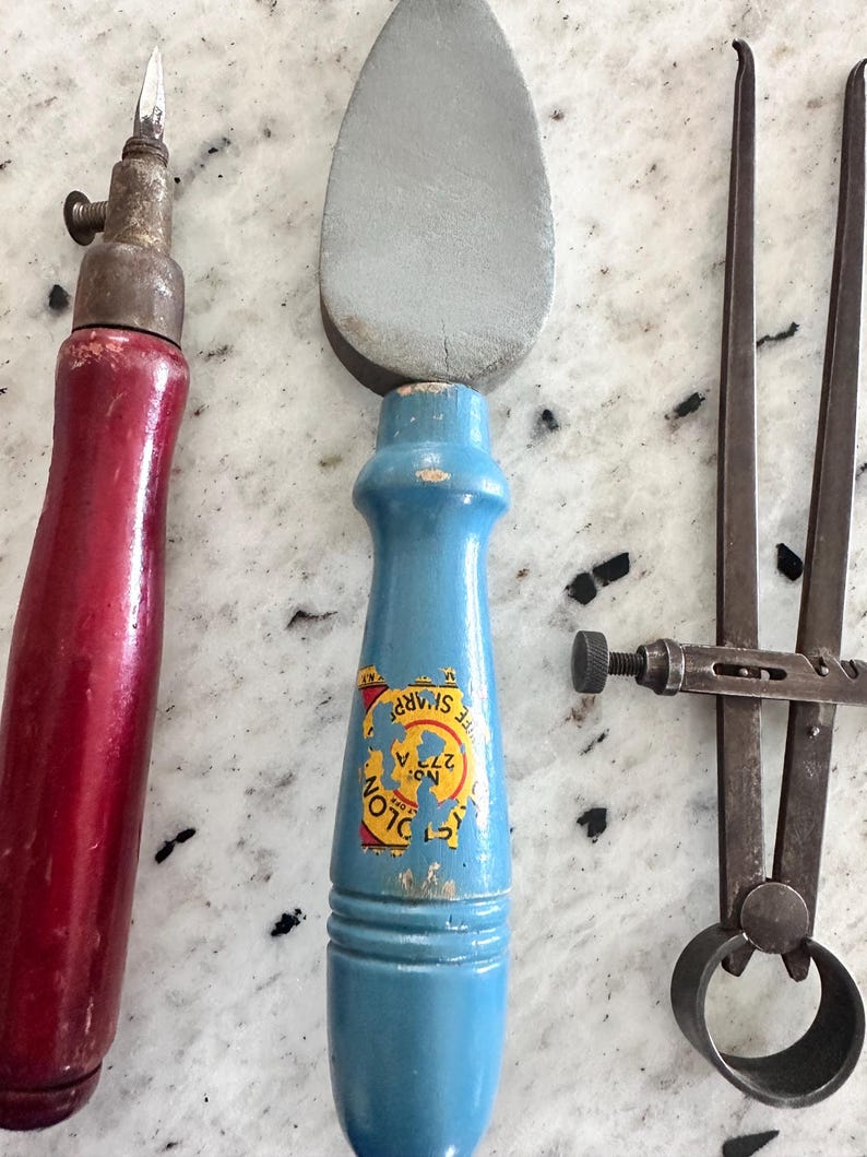 May include: A set of three tools including a red-handled awl, a blue-handled sharpening stone with a label reading Cyclone No. 272 A, and a metal caliper. The tools feature worn wooden handles and metallic components, showing signs of age and use.