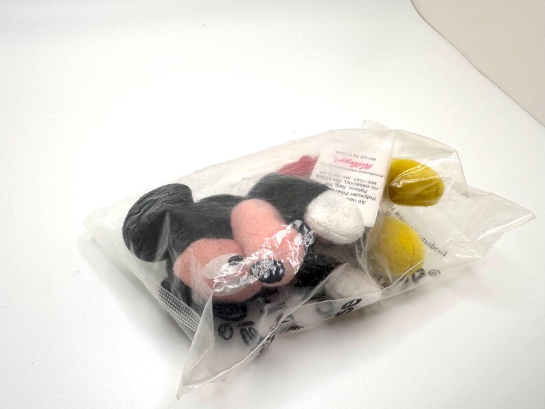 Pu&ograve; includere: Un sacchetto di plastica trasparente contenente diverse parti in peluche di Topolino. Le parti includono orecchie nere, viso e mani rosa, guanti bianchi e cerchi gialli. Il sacchetto &egrave; sigillato.