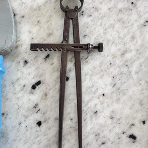 May include: A dark metal caliper tool featuring two long, straight legs connected by a circular pivot at the top. A horizontal bar with a notched, gear-like edge and a small threaded adjustment knob is positioned across the center of the legs.