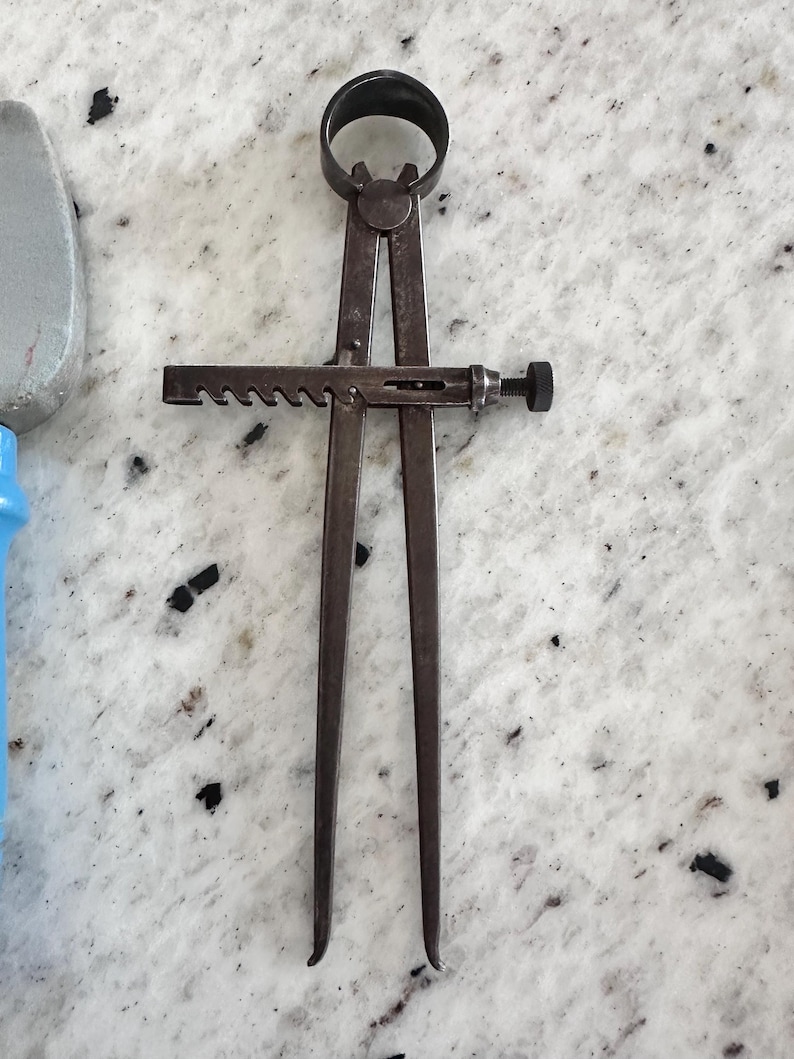 May include: A dark metal caliper tool featuring two long, straight legs connected by a circular pivot at the top. A horizontal bar with a notched, gear-like edge and a small threaded adjustment knob is positioned across the center of the legs.