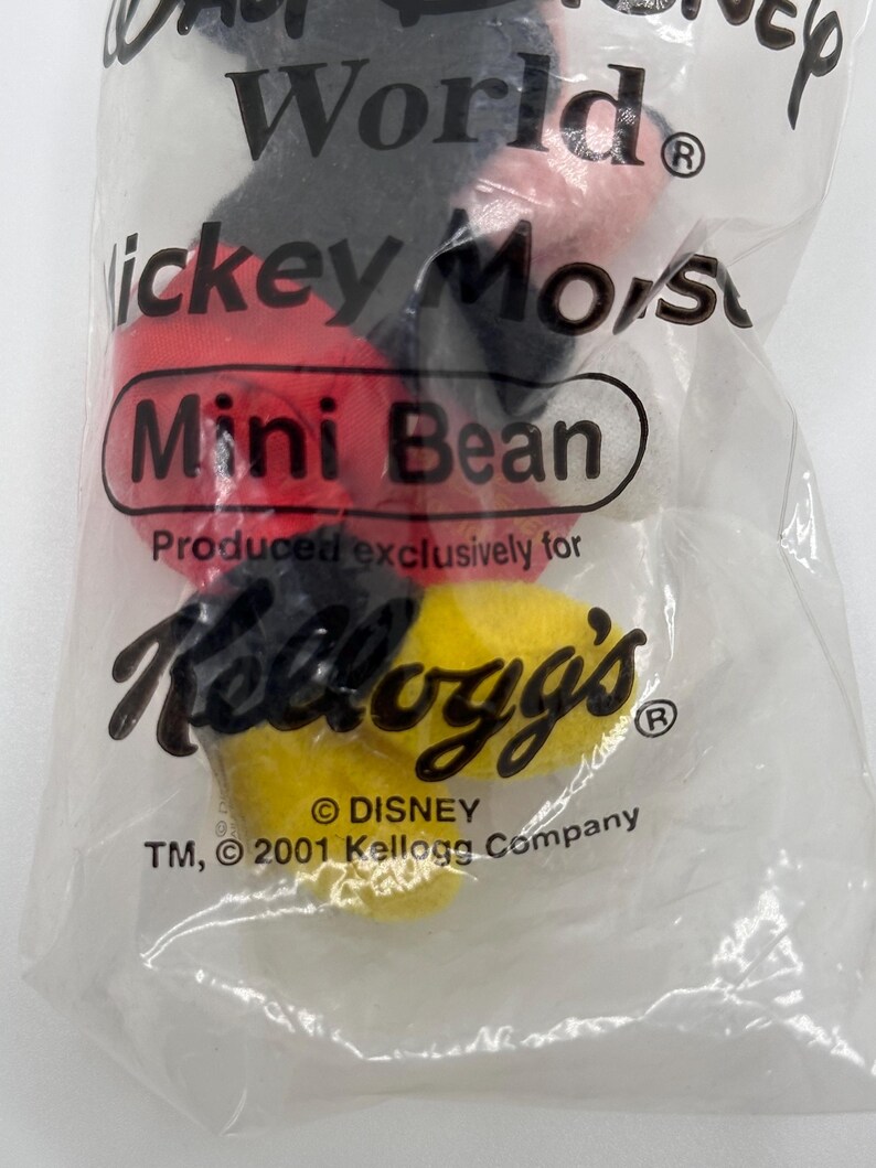 Pu&ograve; includere: Un sacchetto di plastica trasparente contenente un peluche Mickey Mouse Mini Bean. La borsa presenta il testo "World", "Mickey Mouse" e "Kellogg's". Il giocattolo ha elementi rossi, neri e gialli. La borsa &egrave; contrassegnata dai marchi Disney e Kellogg's.