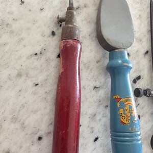 May include: A tool with a red wooden handle and a metal tip with a screw, alongside a tool with a blue wooden handle and a gray stone head. The blue handle features a yellow label with the text "SHARP" and "COLON" partially visible.