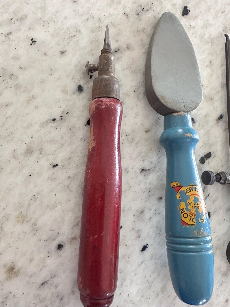 May include: A tool with a red wooden handle and a metal tip with a screw, alongside a tool with a blue wooden handle and a gray stone head. The blue handle features a yellow label with the text "SHARP" and "COLON" partially visible.