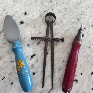 May include: A set of three vintage hand tools including a putty knife with a blue wooden handle featuring a yellow logo, a metal divider caliper with a rack and pinion adjustment mechanism, and a small awl with a dark red wooden handle.