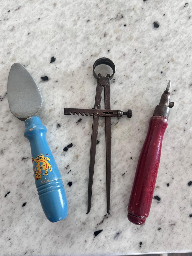May include: A set of three vintage hand tools including a putty knife with a blue wooden handle featuring a yellow logo, a metal divider caliper with a rack and pinion adjustment mechanism, and a small awl with a dark red wooden handle.