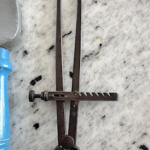 May include: A dark metal caliper tool featuring two long, pointed arms connected by a circular pivot base. A horizontal metal bar with a series of notches and a small adjustment screw is attached to the arms to lock the width of the opening.