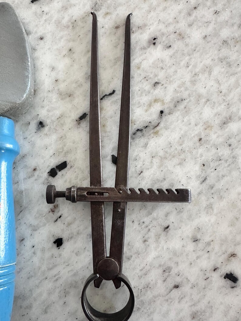 May include: A dark metal caliper tool featuring two long, pointed arms connected by a circular pivot base. A horizontal metal bar with a series of notches and a small adjustment screw is attached to the arms to lock the width of the opening.