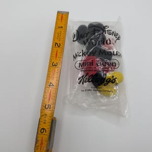 Pu&ograve; includere: Un oggetto da collezione Mickey Mouse Mini Bean non aperto in un sacchetto di plastica trasparente. La borsa presenta i loghi Disney e Kellogg's, con testo che indica che &egrave; stato prodotto da Kellogg's nel 2001. Un righello di legno &egrave; accanto alla borsa.
