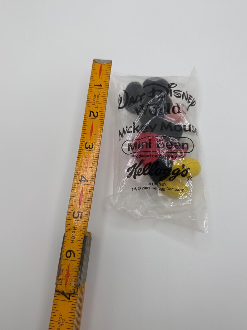 Pu&ograve; includere: Un oggetto da collezione Mickey Mouse Mini Bean non aperto in un sacchetto di plastica trasparente. La borsa presenta i loghi Disney e Kellogg's, con testo che indica che &egrave; stato prodotto da Kellogg's nel 2001. Un righello di legno &egrave; accanto alla borsa.