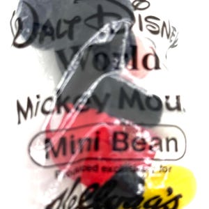 Pu&ograve; includere: Una confezione di plastica trasparente contenente un giocattolo Mickey Mouse Mini Bean. La confezione presenta il testo "Walt Disney World" e "Kellogg's". Il giocattolo di Mickey Mouse ha colori nero, rosso e giallo.