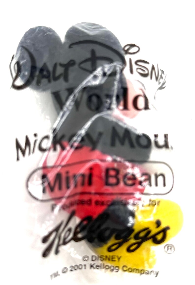 Pu&ograve; includere: Una confezione di plastica trasparente contenente un giocattolo Mickey Mouse Mini Bean. La confezione presenta il testo "Walt Disney World" e "Kellogg's". Il giocattolo di Mickey Mouse ha colori nero, rosso e giallo.