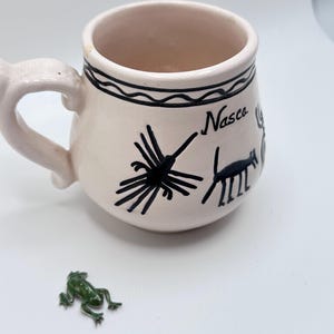 May include: A cream-colored ceramic mug featuring black hand-painted line drawings of a spider and a four-legged animal, with the text "Nasca" written in black script. A small, dark green frog figurine is positioned in front of the mug.