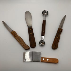May include: A collection of five kitchen utensils, including a small cleaver, a butter spreader, two knives, and a melon baller. The utensils have wooden handles and stainless steel blades. The cleaver measures approximately 5 inches in length.