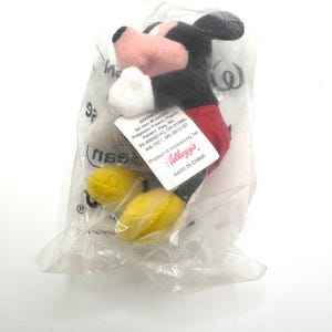 Pu&ograve; includere: Un piccolo peluche di Topolino in un sacchetto di plastica trasparente. Il giocattolo ha dettagli neri, rossi, bianchi, rosa e gialli. Un'etichetta con testo &egrave; attaccata. La confezione include il logo Kellogg's.